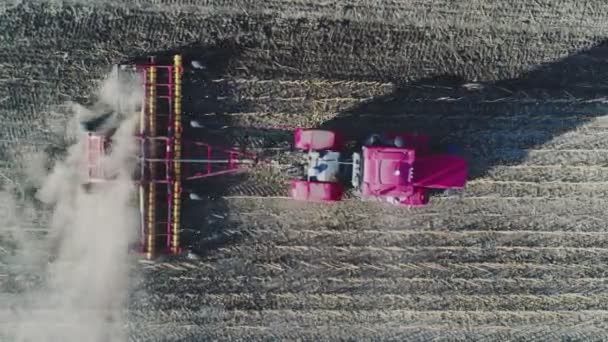 Vue aérienne d'un tracteur rouge avec système de herse labourant sur un champ agricole cultivé, pilier de pistes de poussière derrière, préparation du sol pour la plantation de nouvelles cultures — Video