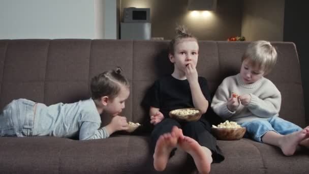 Tiga saudara duduk di sofa dan makan popcorn di rumah — Stok Video