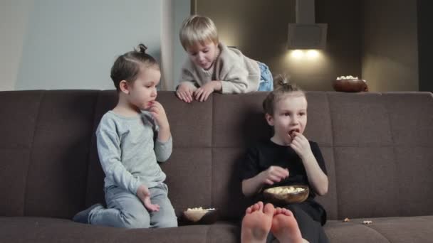 Trzech braci siedzi na kanapie i je popcorn oglądając telewizję w domu. — Wideo stockowe