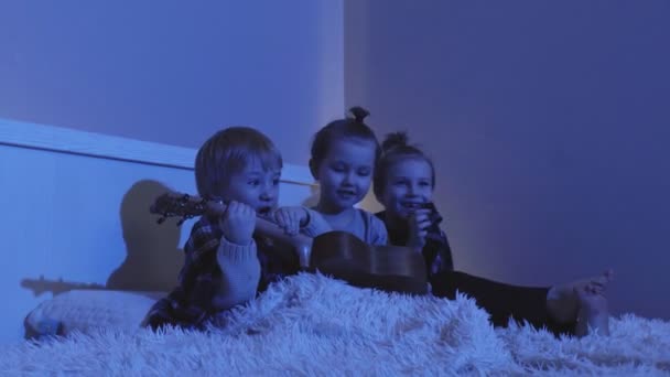 Τρία αγοράκια κάθονται στο κρεβάτι και παίζουν κιθάρα στο δωμάτιο τη νύχτα. — Αρχείο Βίντεο