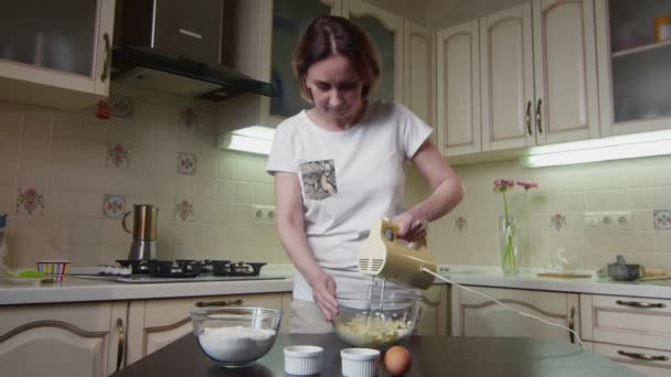 Ama de casa mujer utiliza mezclador para mezclar ingredientes para pastel en la cocina — Vídeos de Stock