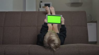 Siyah kapüşonlu şirin çocuk kanepeye uzanıyor ve yeşil ekranlı tablet kullanıyor.