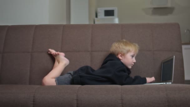 Lindo niño en negro sudadera con capucha pone en sofá y utiliza portátil — Vídeo de stock