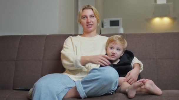 Młoda matka siedzi z synem na kanapie i ogląda telewizję — Wideo stockowe