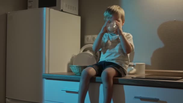 El niño se sienta en la mesa de la cocina y bebe leche por la noche. — Vídeo de stock
