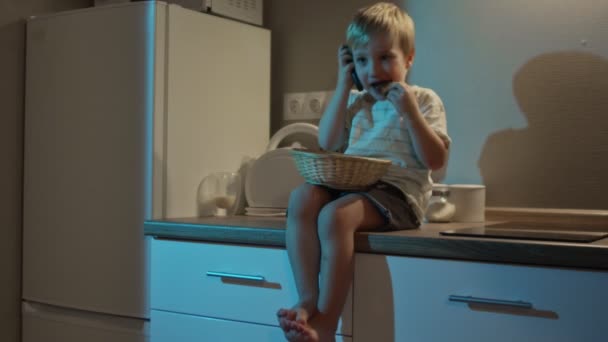 Niño feliz se sienta en la mesa de la cocina, come galletas y habla por teléfono por la noche — Vídeos de Stock