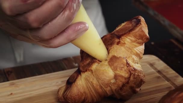 Närbild skjuta en manlig konditor fyller en croissant med grädde fyllning med en bakelse spruta — Stockvideo