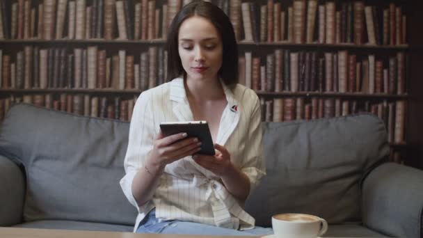 Het meisje zit in de bibliotheek en leest een e-book. — Stockvideo