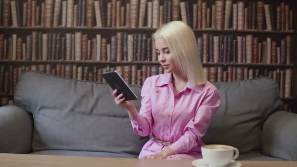 Дівчина сидить у бібліотеці і читає електронну книгу — стокове відео