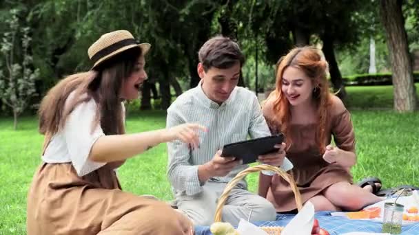 Facet gra w grę na urządzeniu, a dziewczyny obok niego wspierają go w przyrodzie latem — Wideo stockowe