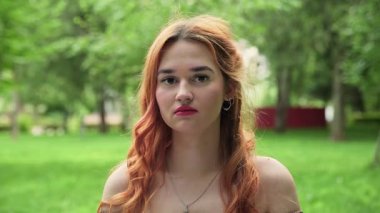 Ciddi yüzlü güzel kızıl saçlı bir kız yazın doğada kameraya bakar.