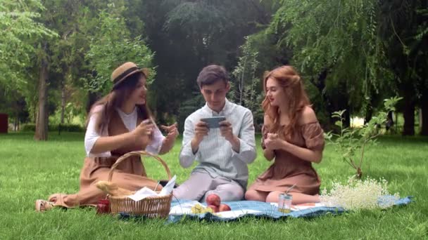 El chico juega el juego en el dispositivo, y las chicas a su lado lo apoyan en la naturaleza en el verano — Vídeos de Stock