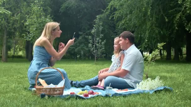 Женщина фотографирует ребенка и мужчину на летнем пикнике на природе по телефону — стоковое видео