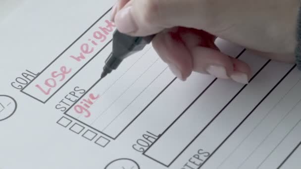 Una mujer de cerca, con un bolígrafo rojo, escribe los escalones de su meta en un primer plano de cuaderno — Vídeo de stock