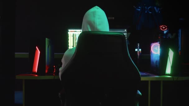Portret tajemniczego hakera w kapturze, siedzącego przy stole i patrzącego w kamerę po włamaniu do bazy danych w ciemnym pokoju z animacją kodów systemowych w tle. — Wideo stockowe