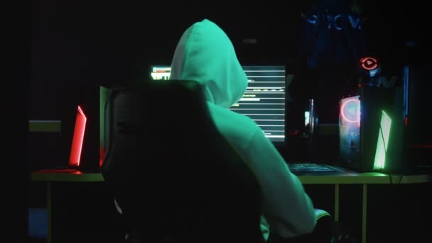 Porträt eines mysteriösen Hackers mit Kapuze, der an einem Tisch sitzt und in die Kamera blickt, nachdem er in einem dunklen Raum eine Datenbank gehackt hat, mit Animation von Systemcodes im Hintergrund. — Stockvideo