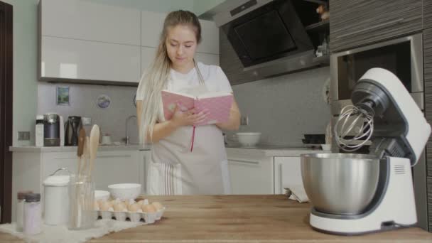 Uma menina na cozinha está à procura de receitas em um livro de receitas ou em um caderno — Vídeo de Stock