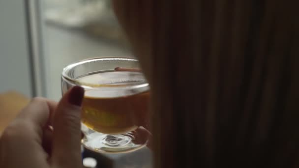 Una joven está sentada cerca de la ventana y sosteniendo una taza de vidrio con té caliente soplando aire fresco en su taza — Vídeos de Stock