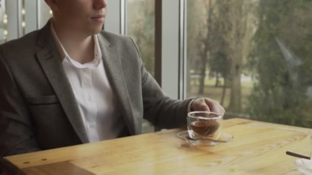 Een zakenman zit bij het raam, heft een glas met groene thee en drinkt het op. — Stockvideo