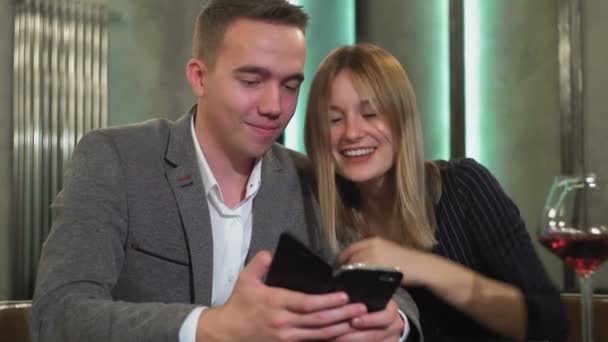 Una pareja atractiva está sentada en una mesa usando un teléfono inteligente, navegando por la web, discutiendo compras en línea, pidiendo comida de un restaurante para una cena romántica — Vídeo de stock