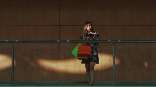 Uma menina asiática em um vestido está de pé com compras de uma loja e está navegando nas páginas da web em seu telefone — Vídeo de Stock