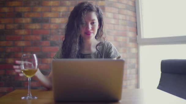 Een jonge vrouw met krullen aan een bureau zit achter een computer en drinkt witte wijn in een glas. — Stockvideo