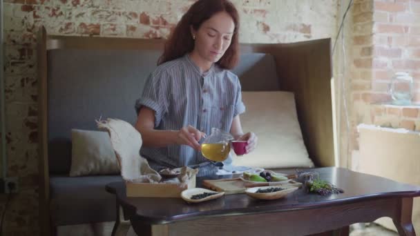 La chica está sentada en la mesa y vierte té verde en una taza de té y lo prueba — Vídeos de Stock