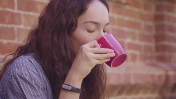 O fată frumoasă cu părul creț bea ceai și se bucură de gustul său pe stradă — Videoclip de stoc