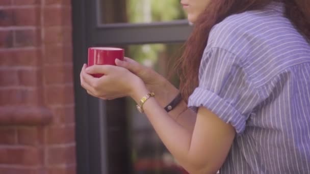Una hermosa chica de pelo rizado bebe té y disfruta de su sabor en la calle — Vídeos de Stock