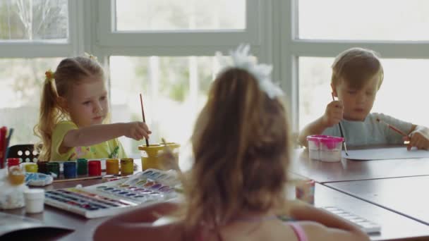 Los niños se sientan en la mesa y dibujan sobre papel con diferentes colores y pinceles — Vídeo de stock