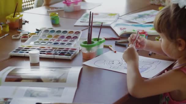 Yan bakışta küçük bir kız masada oturuyor ve farklı renkler ve fırçalarla kağıda resim çiziyor. — Stok video