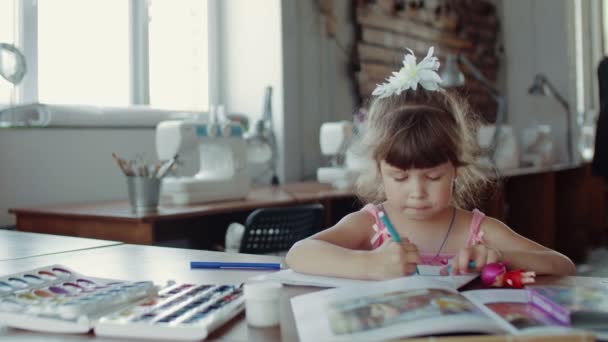 Ein kleines Mädchen sitzt an einem Tisch und zeichnet auf Papier mit verschiedenen Farben und Pinseln — Stockvideo