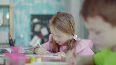Yan bakışta küçük bir kız masada oturuyor ve farklı renkler ve fırçalarla kağıda resim çiziyor.