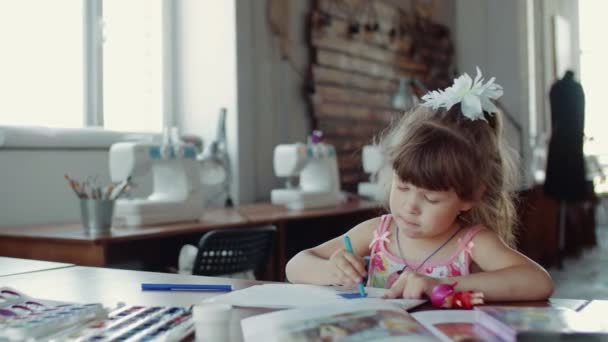 Ein kleines Mädchen sitzt an einem Tisch und zeichnet auf Papier mit verschiedenen Farben und Pinseln — Stockvideo