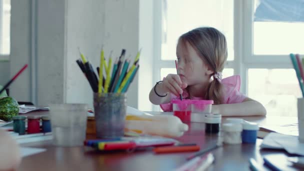 Ein kleines Mädchen sitzt an einem Tisch und zeichnet auf Papier mit verschiedenen Farben und Pinseln — Stockvideo