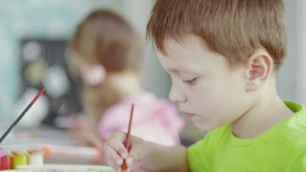 Vista lateral un niño y una niña están sentados en una mesa y dibujando sobre papel con diferentes colores y pinceles — Vídeo de stock