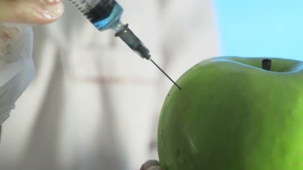 Primo piano di un umano con un cappotto medico e guanti che inietta una siringa in una mela con del liquido su sfondo blu — Video Stock