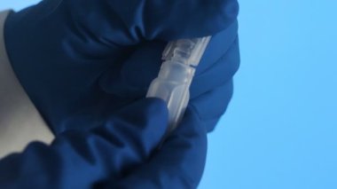 Mavi arka planda plastik bir ampul aşıyı açan lateks eldivenli bir elin yakın çekimi.