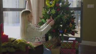 Süveter giyen genç ve güzel bir sarışın Noel ağacının yanında oturuyor ve bir Noel balosu düzenliyor. Evin penceresinin yanında altında hediyeler olan bir Noel ağacı..