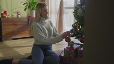 Süveter giyen genç ve güzel bir sarışın Noel ağacının yanında oturuyor ve bir Noel balosu düzenliyor. Evin penceresinin yanında altında hediyeler olan bir Noel ağacı..