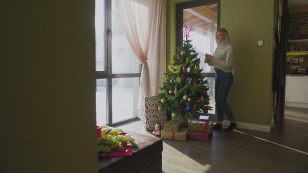 Uma linda jovem loira em uma camisola se aproxima da árvore de Natal e coloca uma bola de Natal. Uma árvore de Natal junto à janela da casa com um monte de presentes debaixo dela. — Vídeo de Stock