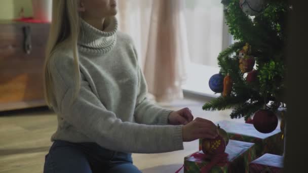 Piękna młoda blondynka w swetrze siedzi obok choinki i zakłada świąteczną piłkę. Choinka przy oknie w domu z kupą prezentów pod spodem.. — Wideo stockowe