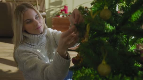 En vacker ung blondin i en tröja sitter bredvid julgranen och sätter på sig en julboll. En julgran vid fönstret i huset med en massa presenter under. — Stockvideo
