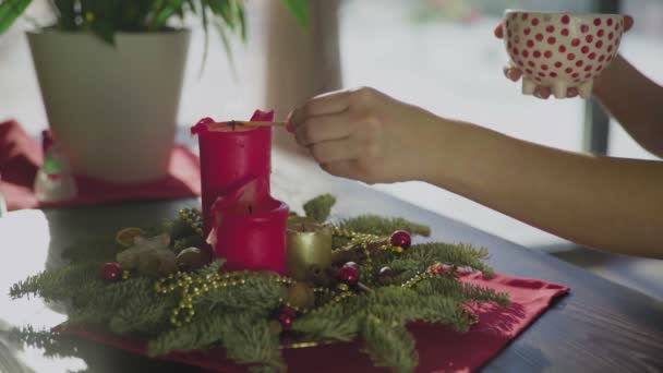 An unrecognizable girl lights an evergreen wreath with two red candles and one golden candle. Christmas traditions and customs of Christians on the eve of the holiday. A symbol of the holiday. — Stock Video