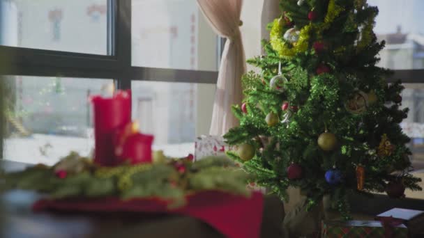 Una corona de hoja perenne con dos velas rojas y una vela dorada. En el fondo, un árbol de Navidad decorado. Las tradiciones y costumbres navideñas de los cristianos en vísperas de la fiesta. — Vídeo de stock