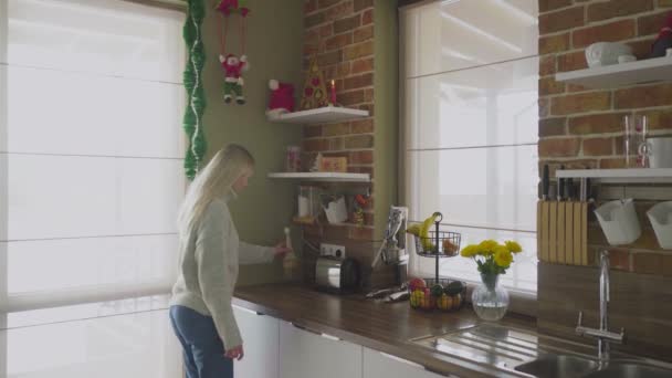 Una joven rubia hermosa entra en la cocina y corrige objetos y varias decoraciones. Navidad — Vídeos de Stock