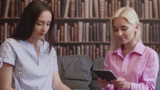 Due ragazze si siedono in biblioteca e leggono un libro in forma elettronica — Video Stock