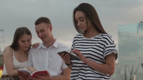 Дві дівчини і хлопець сидять в парку протягом дня читаючи електронну книгу — стокове відео