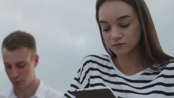 Två tjejer och en kille som sitter i parken under dagen och läser en e-bok — Stockvideo