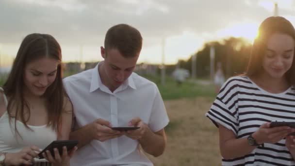 Två tjejer och en kille som sitter i parken under dagen och läser en e-bok — Stockvideo